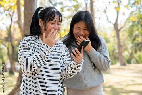 Two cheerful female best friends are laughing, watching something funny on a smartphone in a park.