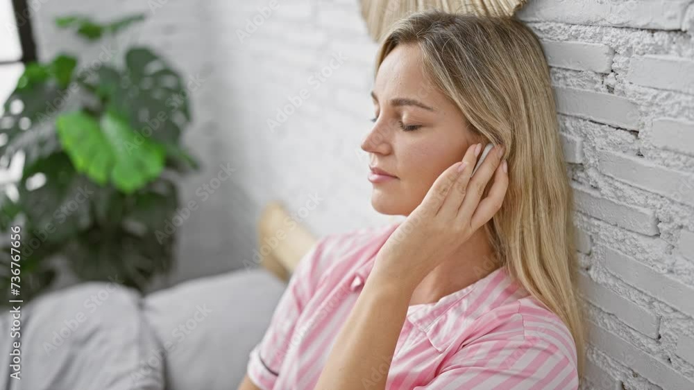 A serene young blonde woman enjoys wireless technology in her modern white-brick bedroom. A serene young blonde woman enjoys wireless technology in her modern white-brick bedroom.