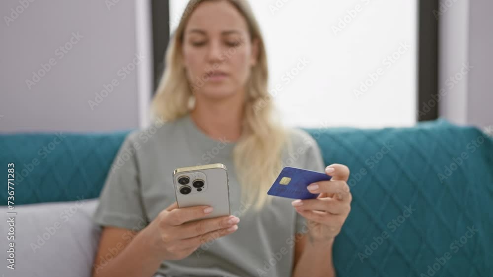 A pensive young woman examines a credit card while holding a smartphone in a modern living room. A pensive young woman examines a credit card while holding a smartphone in a modern living room.