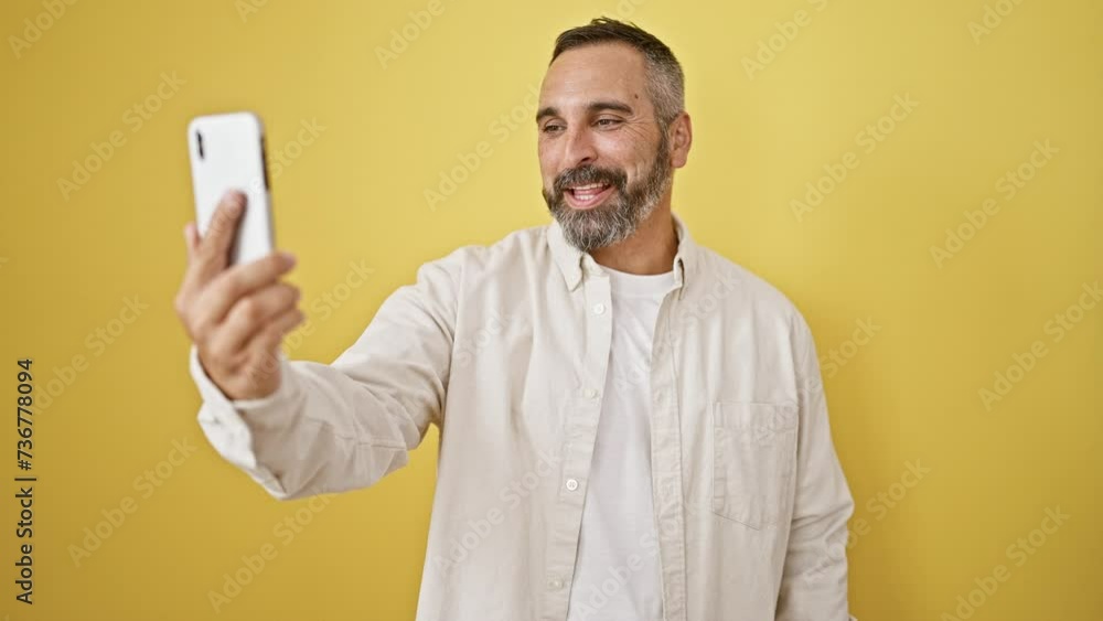 Smiling bearded man taking selfie with phone against yellow background Smiling bearded man taking selfie with phone against yellow background