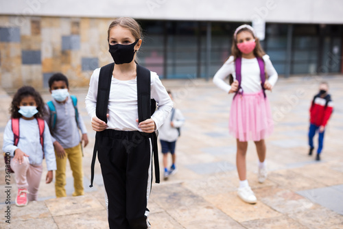 Portrait of schoolgirl in medical mask standing on the street, kids on background