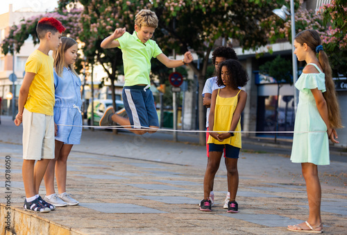Children active summer games. Interested happy friendly tweenagers playing chinese jump rope in flowering city square..