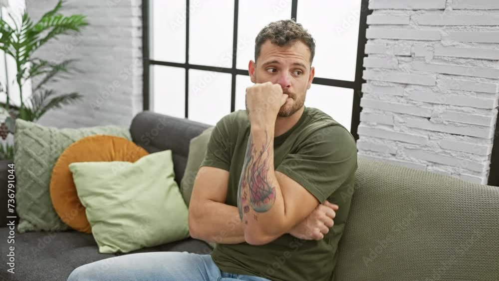 At home, a stressed young man sits on the sofa nervously biting his nails, hands trembling with anxiety. indoor portrait of handsome, fearful, bearded adult grappling with stress problem. At home, a stressed young man sits on the sofa nervously biting his nails, hands trembling with anxiety. indoor portrait of handsome, fearful, bearded adult grappling with stress problem.
