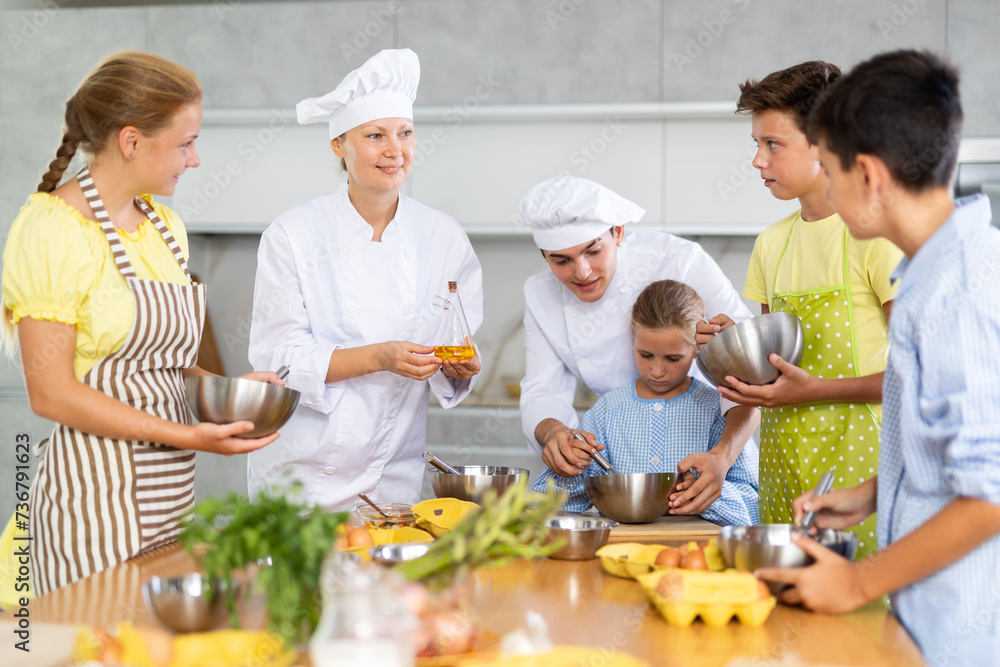 Two experienced chefs, friendly woman and man in white uniform giving culinary lesson to group of inquisitive tweens, teaching basic cooking skills