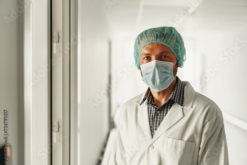 Male doctor in white gown and mask standing in medical clinic and looks camera