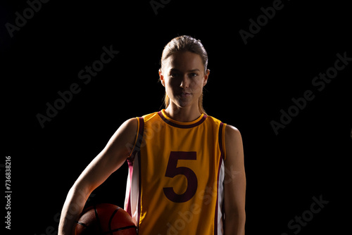 Young Caucasian female basketball player poses confidently in a basketball uniform on a black backgr