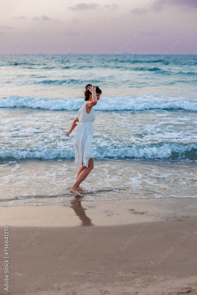 happy young girl in a white summer dress walks along the beach at sunset.