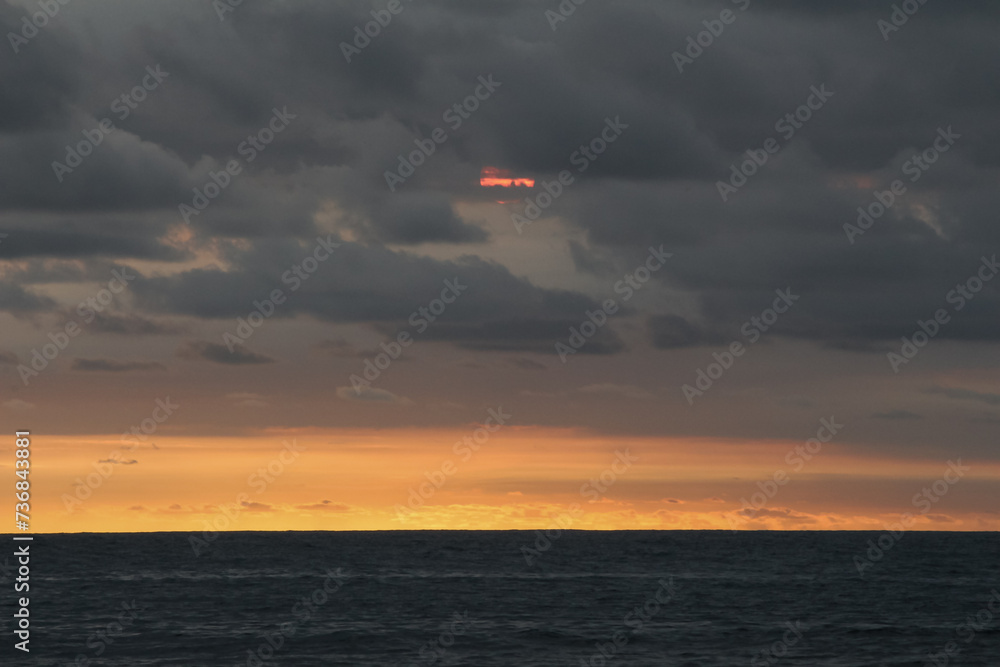 The sun was reddish above the vast ocean, which was covered in black clouds, a sign of cloudiness. Part of the sky was orange at the end of the ocean and part of it was covered by black clouds. The sun was reddish above the vast ocean, which was covered in black clouds, a sign of cloudiness. Part of the sky was orange at the end of the ocean and part of it was covered by black clouds.