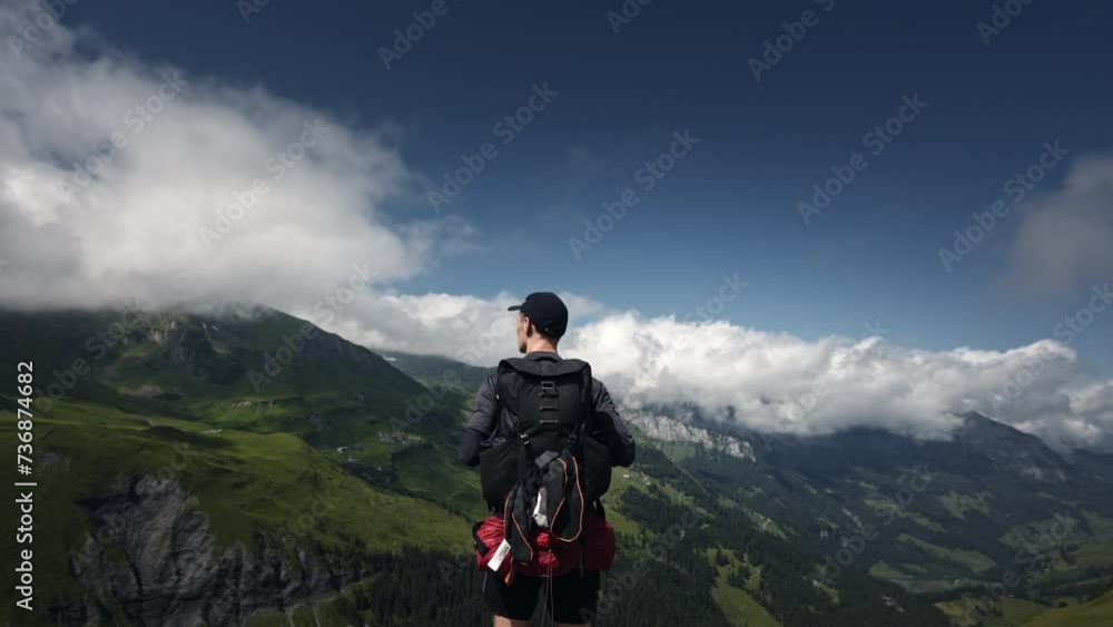 Caucasian male backpacker standing on top of a hill with green valley, mountains and clouds in the background Caucasian male backpacker standing on top of a hill with green valley, mountains and clouds in the background