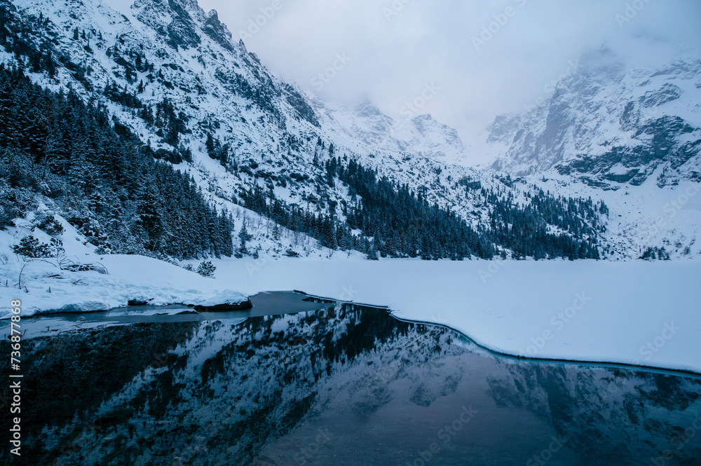 Obraz premium Morskie Oko lake sits in front of a majestic snowy mountain range, creating a stunning natural landscape. The sky is filled with fluffy clouds, enhancing the atmosphere of the scene. Tatry, Poland