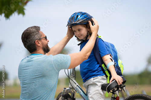 Little kid learning to ride bicycle with father in park. Father teaching son cycling. Father and son learning to ride a bicycle at Fathers day. Father helping his son to wear a cycling helmet.