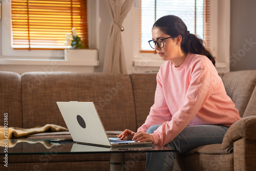 Young Caucasian woman wearing glasses is sitting on couch and using her laptop. Cozy interior. Concept of remote work and freelancing