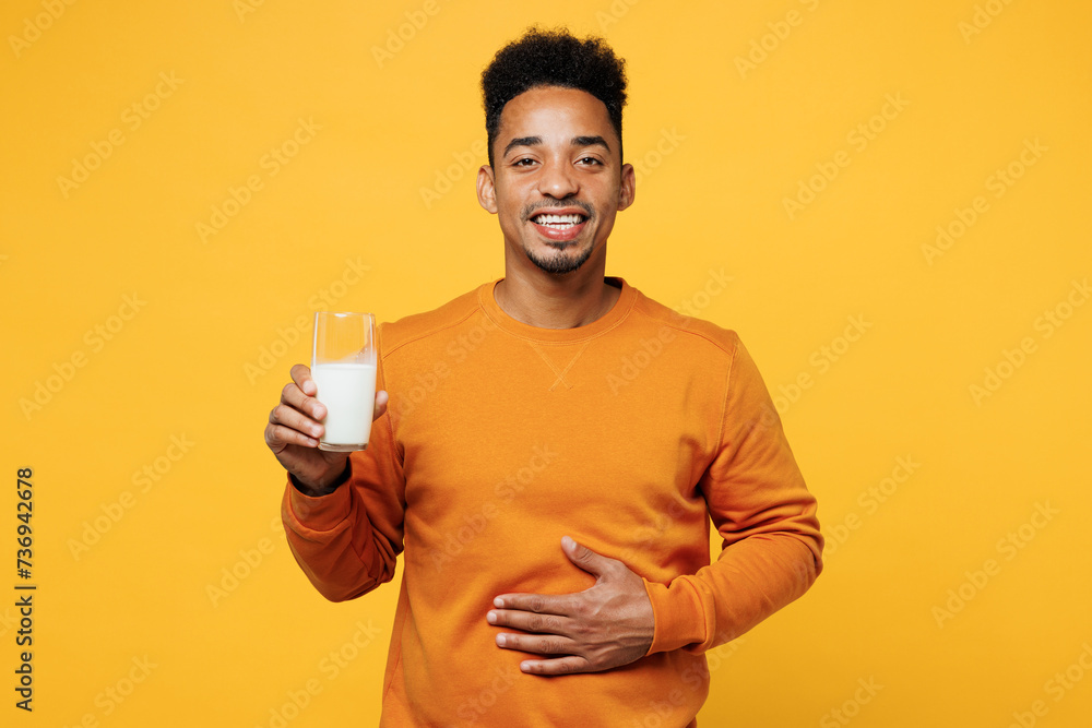 Young man he wear orange sweatshirt casual clothes isolated on plain yellow hold glass of regular milk put hand on belly background studio. Proper nutrition healthy fast food unhealthy choice concept. Young man he wear orange sweatshirt casual clothes isolated on plain yellow hold glass of regular milk put hand on belly background studio. Proper nutrition healthy fast food unhealthy choice concept.