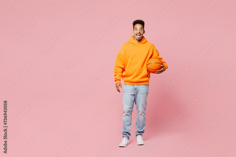 Full body young man of African American ethnicity fan wears yellow hoody casual clothes cheer up support basketball sport team hold in hand ball watch tv live stream isolated on plain pink background. Full body young man of African American ethnicity fan wears yellow hoody casual clothes cheer up support basketball sport team hold in hand ball watch tv live stream isolated on plain pink background.