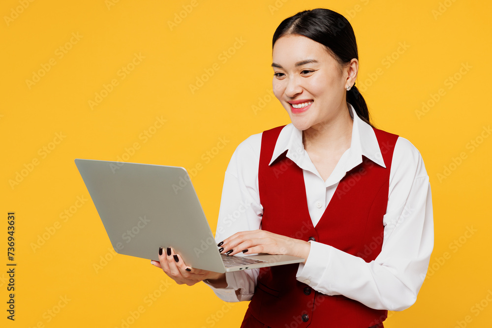 Young smiling corporate lawyer employee business IT woman of Asian ethnicity wear formal red vest shirt work at office hold use laptop pc computer isolated on plain yellow background. Career concept,. Young smiling corporate lawyer employee business IT woman of Asian ethnicity wear formal red vest shirt work at office hold use laptop pc computer isolated on plain yellow background. Career concept,.