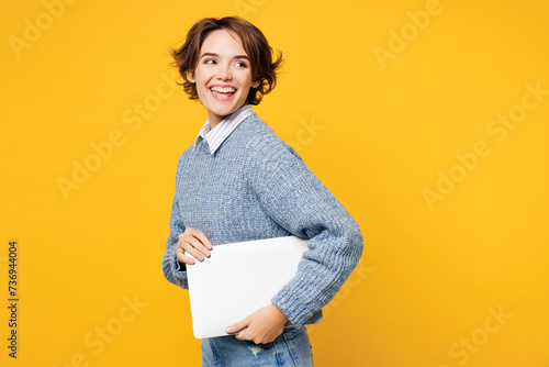Side view young programmer IT woman she wearing grey knitted sweater shirt casual clothes hold closed laptop pc computer chatting online isolated on plain yellow background studio. Lifestyle concept.