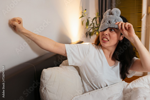 Portrait of angry Caucasian woman removing sleep mask knocking with fists to wall sitting in bed. Insomnia due to noisy neighbors