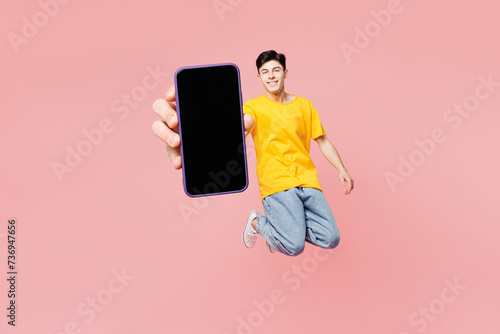 Full body young man he wears yellow t-shirt casual clothes jump high hold in hand show use blank screen area mobile cell phone isolated on plain pastel light pink background studio. Lifestyle concept.