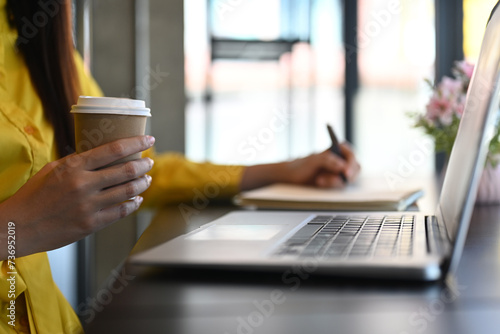 Female office worker holding paper cup of coffee and using laptop on black wooden table