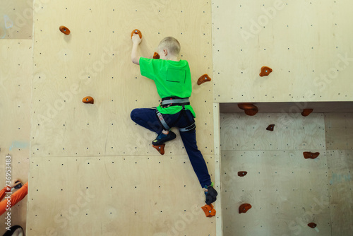 Rock climbing for children. Blond boy in a green shirt is climbing climbing wall. Bouldering class. Artificial track for safe activities and competitions. Mountaineering, safety system, special shoes