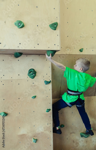 Rock climbing for children. Blond boy in a green shirt is climbing climbing wall. Bouldering class. Artificial track for safe activities and competitions. Mountaineering, safety system, special shoes