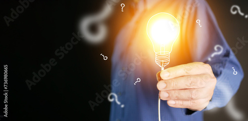 Idea concept. Lightblub is on as a symbol of innovation, inspiration, solution, creativity. Hand holds illuminated light bulb with wire surrounded by question marks. Creative ideas and thinking