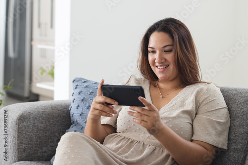 Young plus size biracial woman enjoys gaming on her phone at home, with copy space unaltered photo