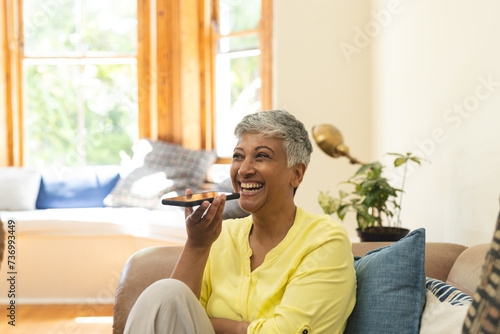 Mature biracial cheerful woman using voice command on smartphone at home photo