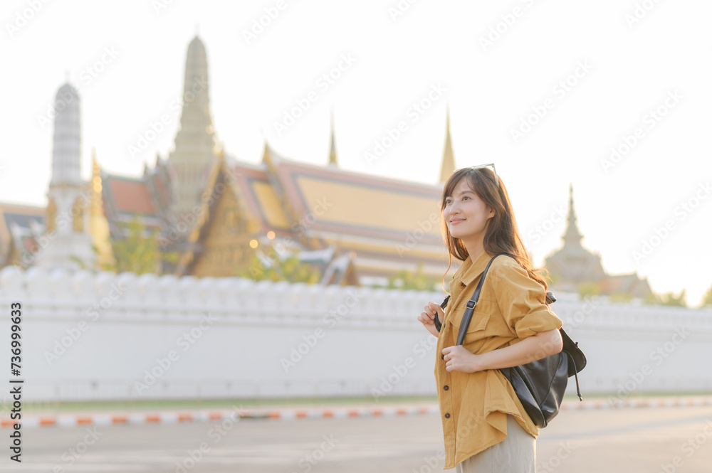 Traveler asian woman in her 30s, backpack slung over her shoulder, explores the intricate details of Wat Pra Kaew with childlike wonder. Sunlight dances on the golden rooftops.