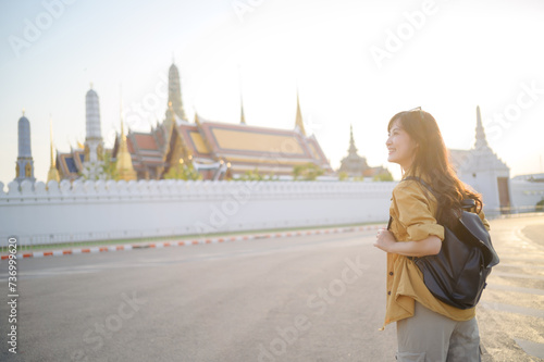 Traveler asian woman in her 30s, backpack slung over her shoulder, explores the intricate details of Wat Pra Kaew with childlike wonder. Sunlight dances on the golden rooftops.