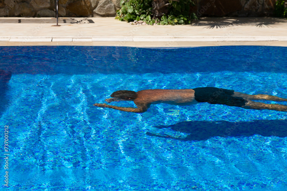 A man in a Hawaiian style swimsuit swims underwater in a pool of blue water. The concept of fun in the pool on vacation.
