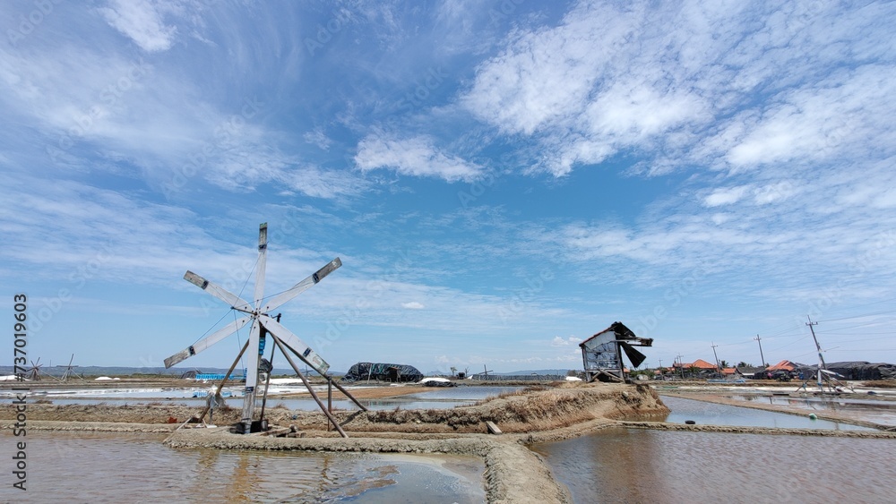 A windmill stands tall on a salt-making land, look a wooden huts as resting places for salt farmers. bright blue sky dotted with white clouds.