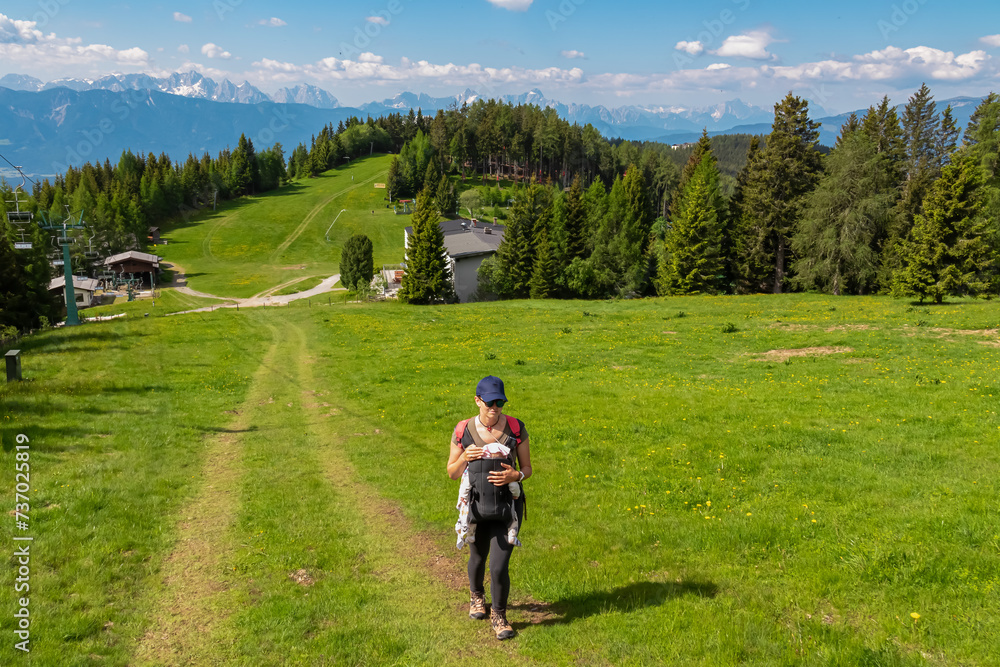 Woman with baby carrier on hiking trail along alpine meadow with scenic view of mountain range Julian Alps seen from Gerlitzen, Carinthia, Austria. Walking next to cable car. Wanderlust Austrian Alps Woman with baby carrier on hiking trail along alpine meadow with scenic view of mountain range Julian Alps seen from Gerlitzen, Carinthia, Austria. Walking next to cable car. Wanderlust Austrian Alps