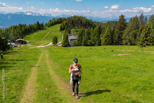 Woman with baby carrier on hiking trail along alpine meadow with scenic view of mountain range Julian Alps seen from Gerlitzen, Carinthia, Austria. Walking next to cable car. Wanderlust Austrian Alps © Chris