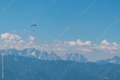 Person paragliding over alpine landscape with panoramic view of majestic mountain range Julian Alps seen from Gerlitzen Alpe, Carnthia, Austria. Dangerous adrenaline sport. Leisure activity in summer