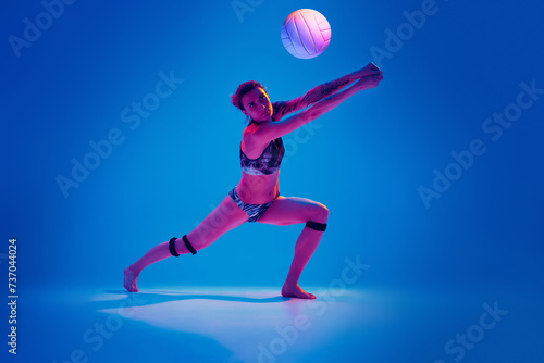 Concentrated athlete woman, beach volleyball player passes ball from below against gradient blue background in pink neon light. Concept of sport, movement, active and healthy lifestyle, power.