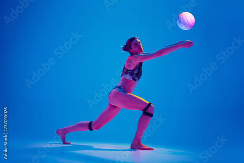 Athlete woman in sportswear playing beach volleyball against gradient blue background in pink neon light. Concept of sport  movement  active and healthy lifestyle  power and strength.