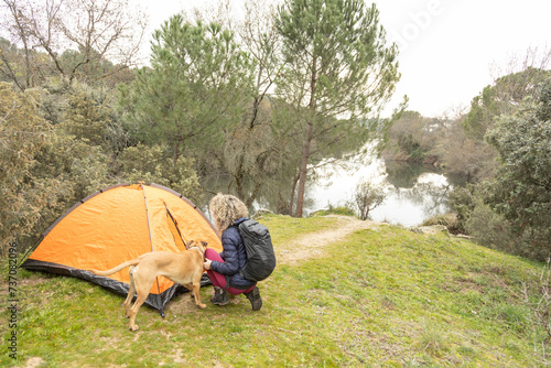 Curly and blonde hair, mountaineer woman and her dog camps with his orange tent