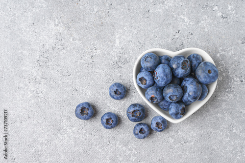 Fresh blueberries and scattering of berries in small white heart shaped bowl on gray concrete background. Organic berries, healthy food, wild berries. Top view, flat lay