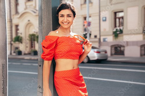 Young beautiful smiling hipster woman in trendy summer red top and skirt clothes. Carefree female posing in the street at sunny day. Positive model outdoors at sunset. Cheerful and happy. Closeup
