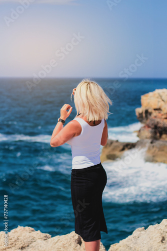 blonde in a white shirt and black skirt stands with her back and looks at the sea on a rocky shore on a sunny day