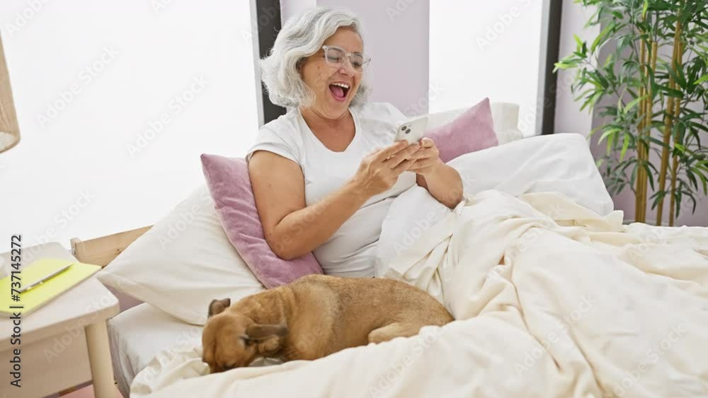 A smiling, grey-haired woman comfortably lounging in bed with her dog, interacting with her smartphone at home. A smiling, grey-haired woman comfortably lounging in bed with her dog, interacting with her smartphone at home.