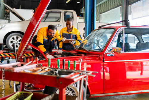 Diversity mechanic teamwork, an Indian man and an African woman in yellow and blue uniforms. A man inspects the car engine with his woman assistant. Automobile repairing service. Vehicle maintenance.