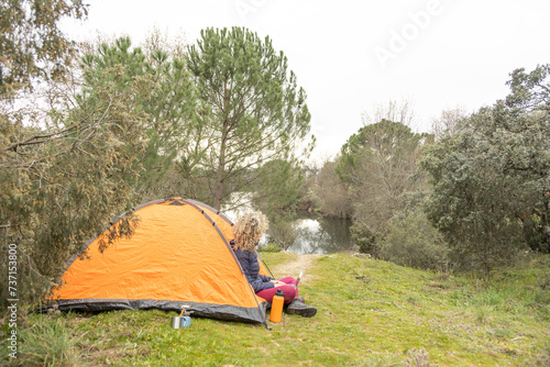 Curly and blonde hair, mountaineer woman camps with his orange tent, teleworking with his laptop