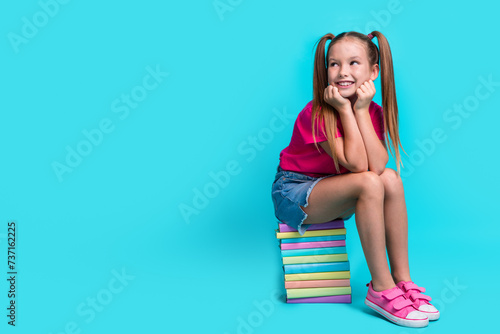 Full length photo of diligent schoolgirl wear t-shirt sit on book look at proposition empty space isolated on teal color background photo