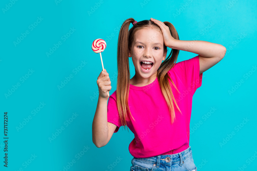 Photo of impressed overjoyed schoolgirl with ponytails wear pink t-shirt hold lollipop arm on head isolated on teal color background