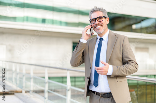 A senior professional in smart business attire is engaged in a pleasant phone conversation, gesturing with one hand as he talks with the modern office building in the background