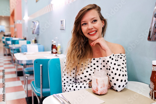 Young beautiful woman sitting at bar drinks a delicious milkshake inside classic vintage american retro bar.