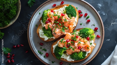 a white plate topped with two pieces of bread covered in salmon and broccoli next to a bowl of pomegranate.