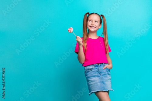 Portrait of satisfied small girl dressed pink t-shirt holding lollipop look at promo empty space isolated on turquoise color background